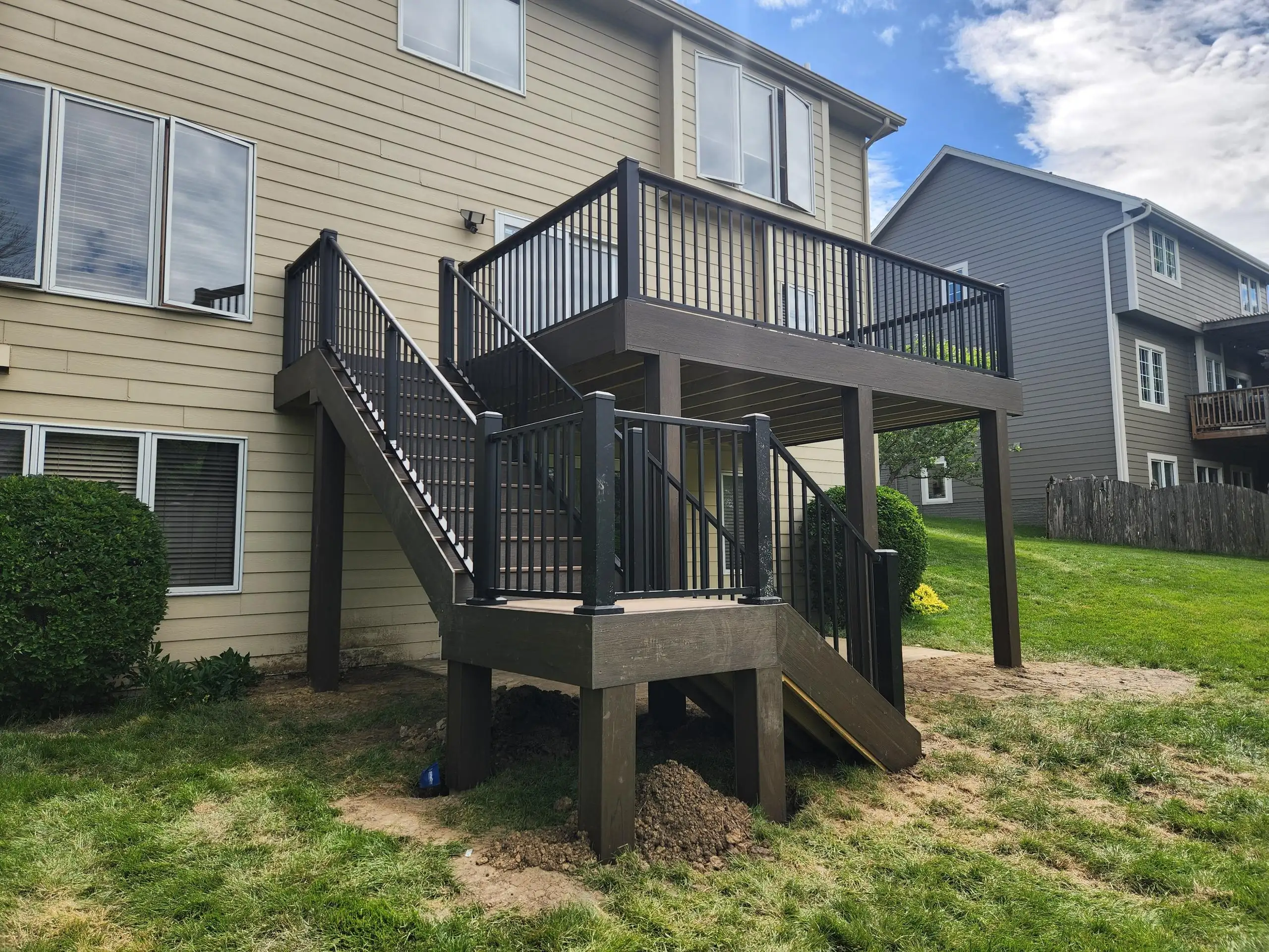 A two-story brown deck with black railings and stairs leads up to the second floor of a tan house.