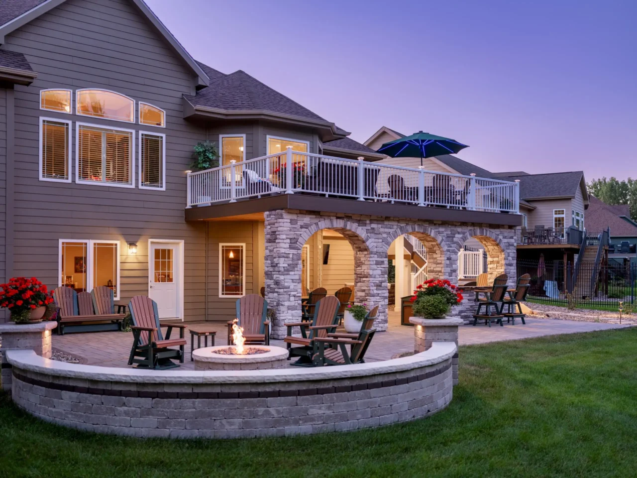 A custom multi-level TimberTech deck and paver patio with a built-in fire pit, stone archways, and deck lighting. This backyard living space in West Des Moines is photographed at dusk.