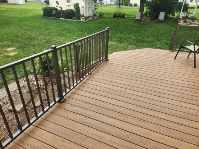 Trex Deck Design Ideas A composite deck. The deck has a light brown color with a wood grain texture and is bordered by dark railings. In the background, a well-maintained green lawn and various plants and shrubs are visible.