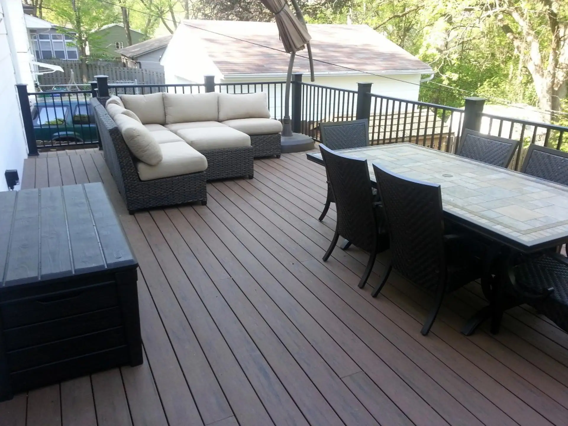 TimberTech vs Trex Composite deck with black railing, furnished with a tan sectional sofa and a tiled dining table with six wicker chairs. A large black storage box is in the foreground.