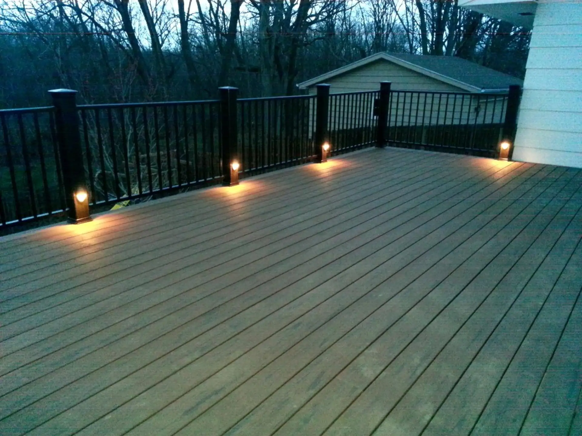 An outdoor composite deck at twilight or night. The deck has a dark railing with square posts, and the posts are illuminated by small, integrated lights near the bottom, casting a warm glow onto the wood-like deck surface.