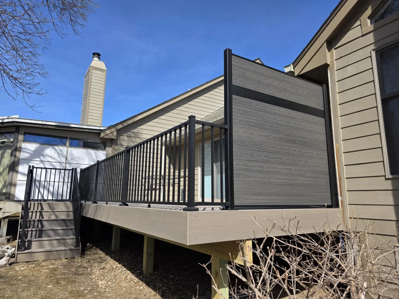 Ahouse with a newly installed outdoor deck. The deck features black metal railings and light brown composite decking. A dark gray privacy screen is mounted to the deck, partially covering a sliding glass door. Stairs lead down from the deck on the left. The house has light tan siding, and a chimney is visible on the roofline against a clear blue sky.
