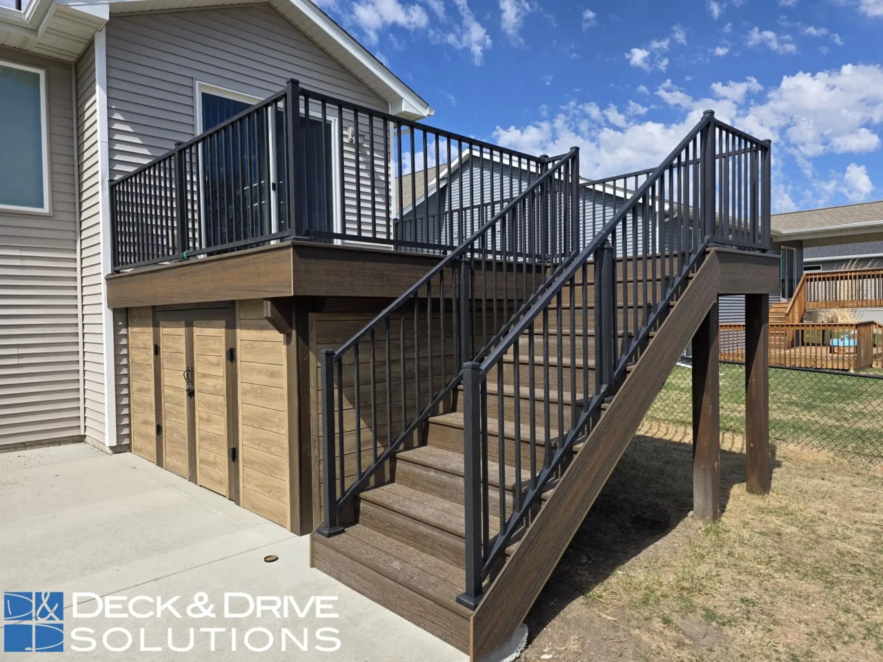A multi-level outdoor deck, featuring dark brown composite decking, black metal railings, and a staircase. The area under the main deck is enclosed by four light-brown, wood-look storage doors. The "DECK and DRIVE SOLUTIONS" logo is visible.