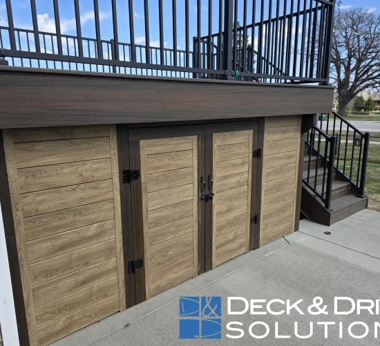 A close-up view of the storage area underneath an elevated dark brown deck. The storage space is enclosed by four light-brown, horizontally planked, wood-look doors with dark-brown trim and black hinges and handles. A set of matching dark brown steps is visible on the right. The logo and text "DECK and DRIVE SOLUTIONS" are positioned in the bottom right corner of the image.